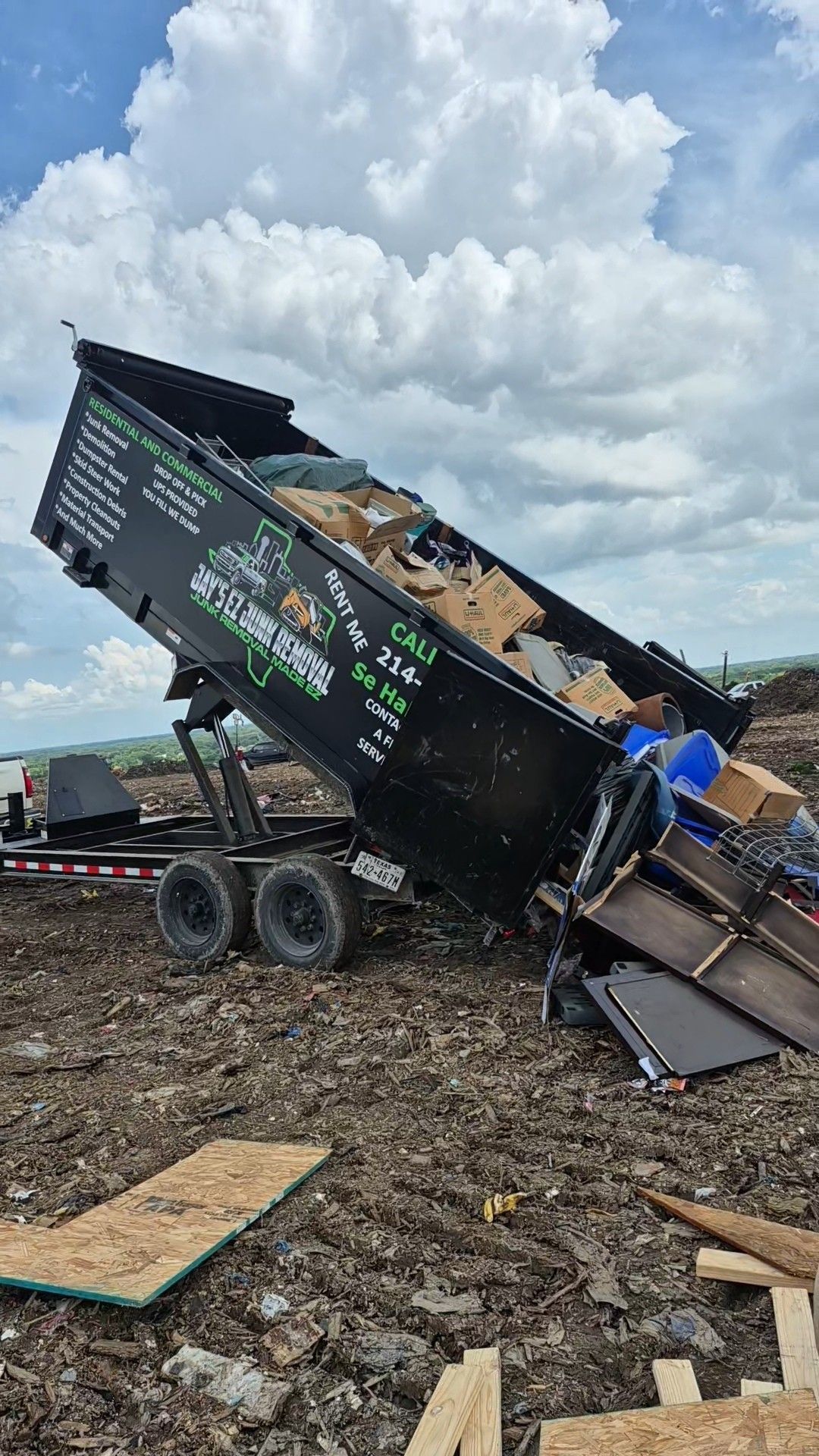 Black dump trailer unloading waste in a muddy field under a cloudy sky.