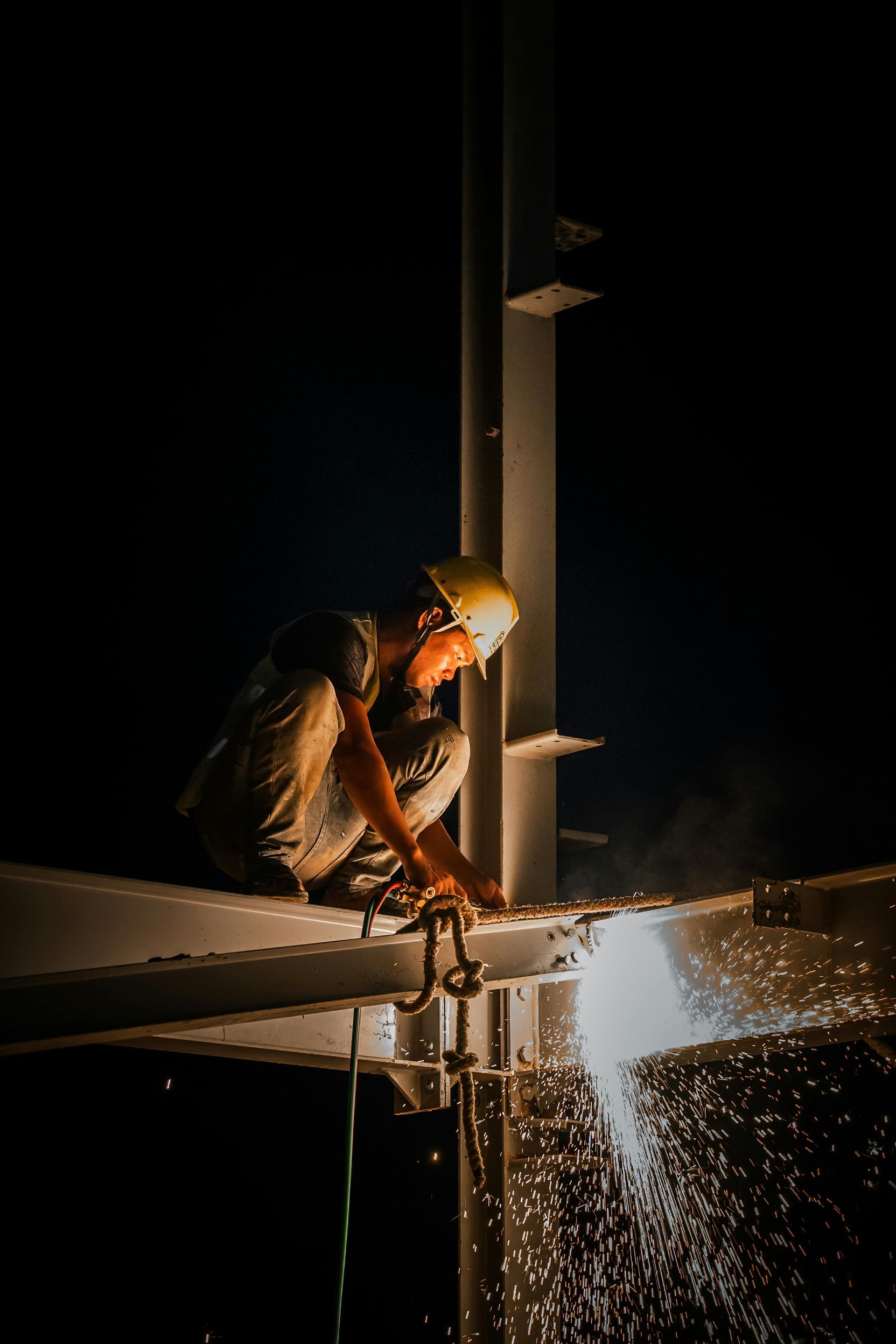 Welder in a yellow hardhat working on metal structure at night, sparks flying.