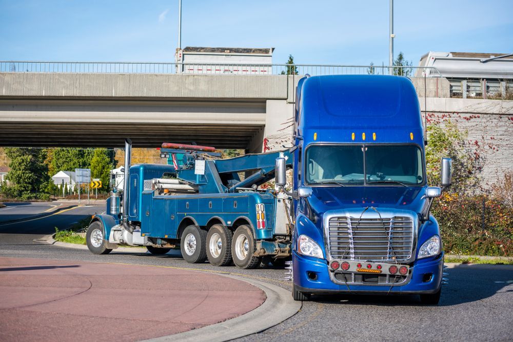 Blue semi-truck being towed by a blue tow truck on a road near a bridge.