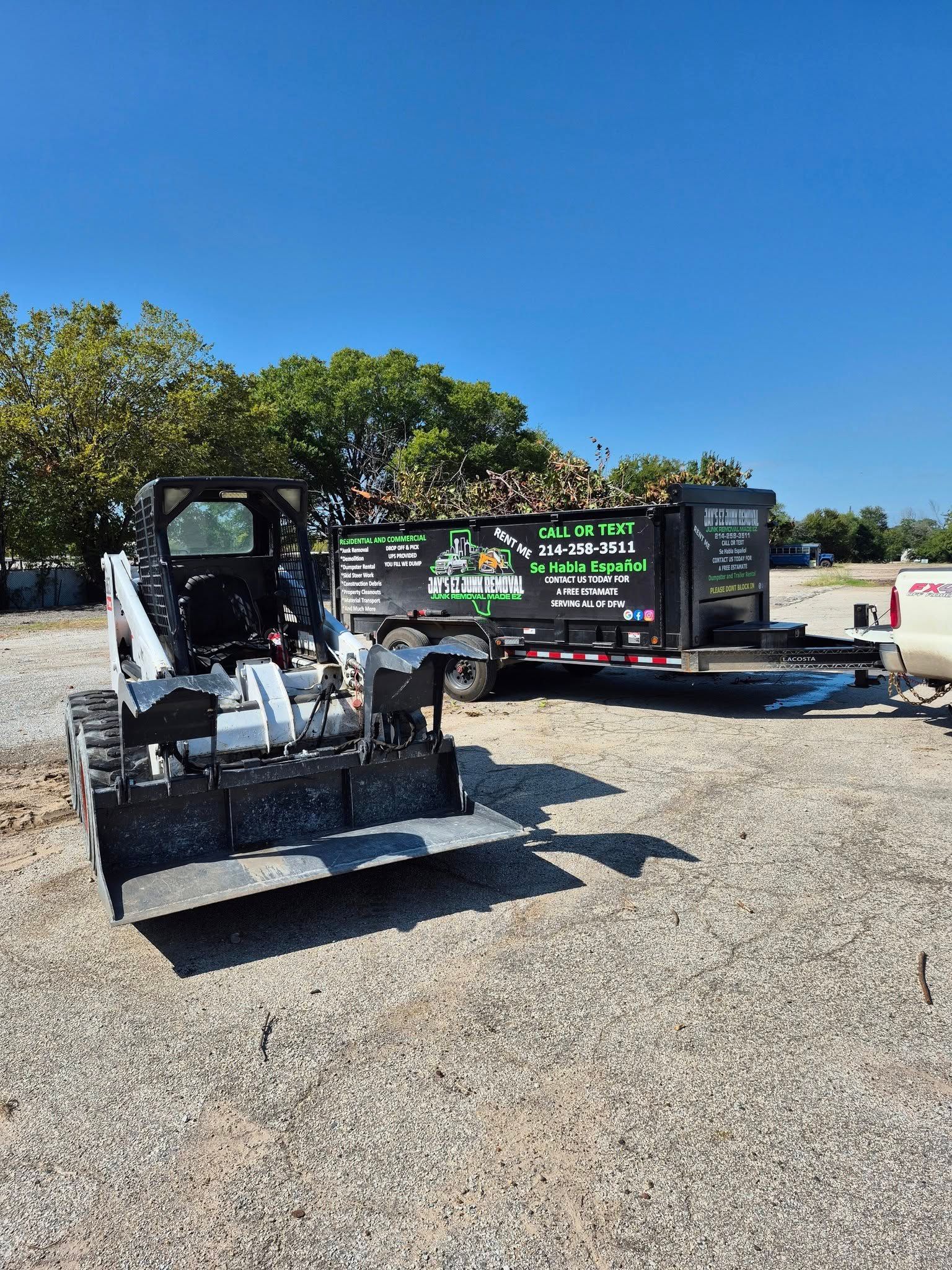 Mini excavator in a gravel lot next to a trailer loaded with wood chips under a blue sky.