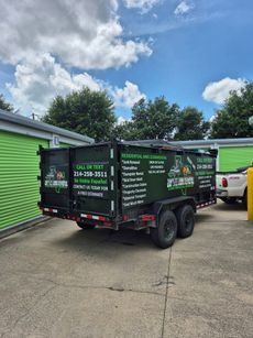 Black trailer with green signage parked next to green storage units. White pickup truck visible. Blue sky, clouds.