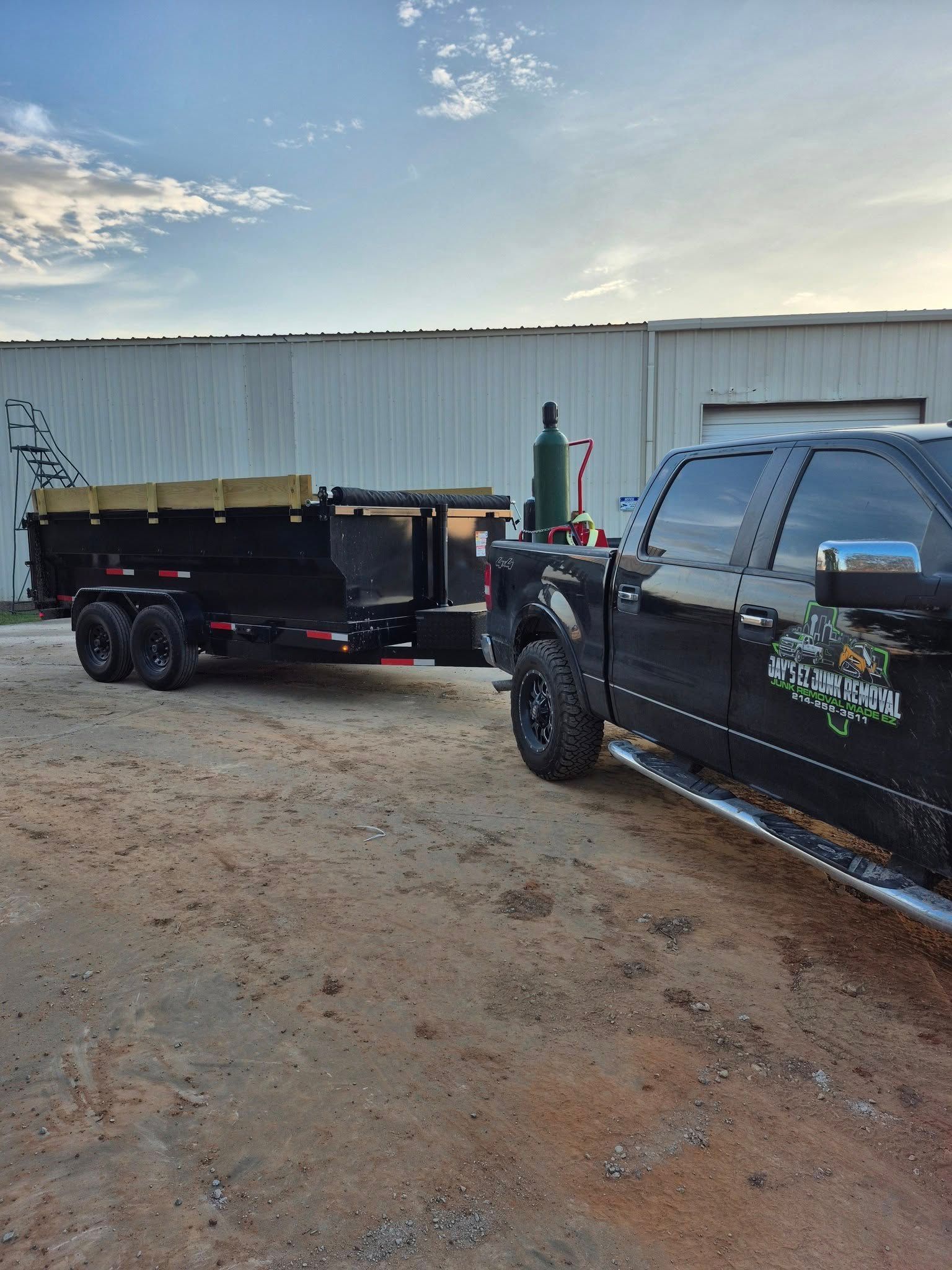 Black truck towing a black dump trailer on dirt, near a white building under a cloudy sky.
