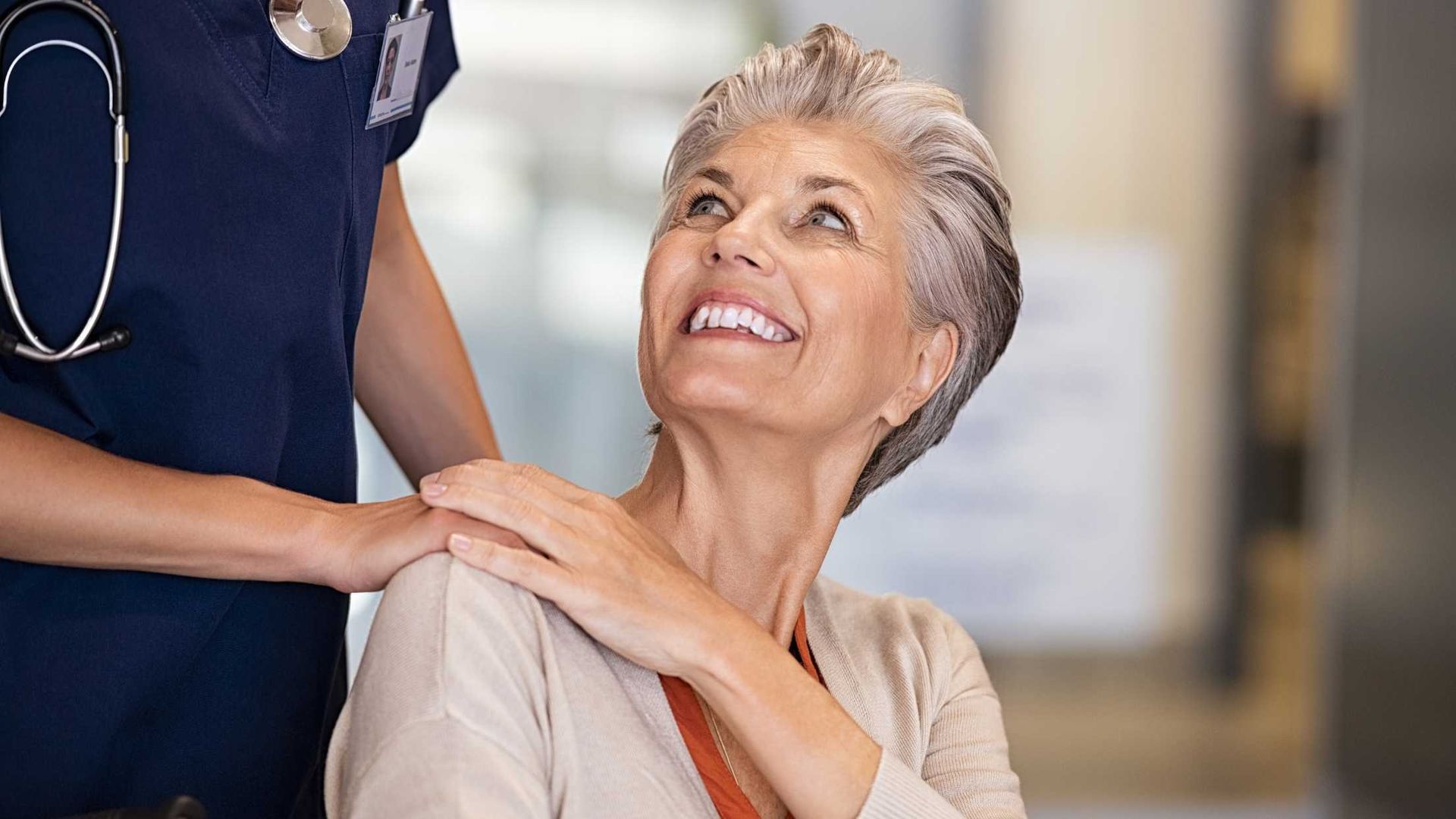 Older woman in wheelchair smiles as a healthcare worker places a hand on her shoulder, looking up.