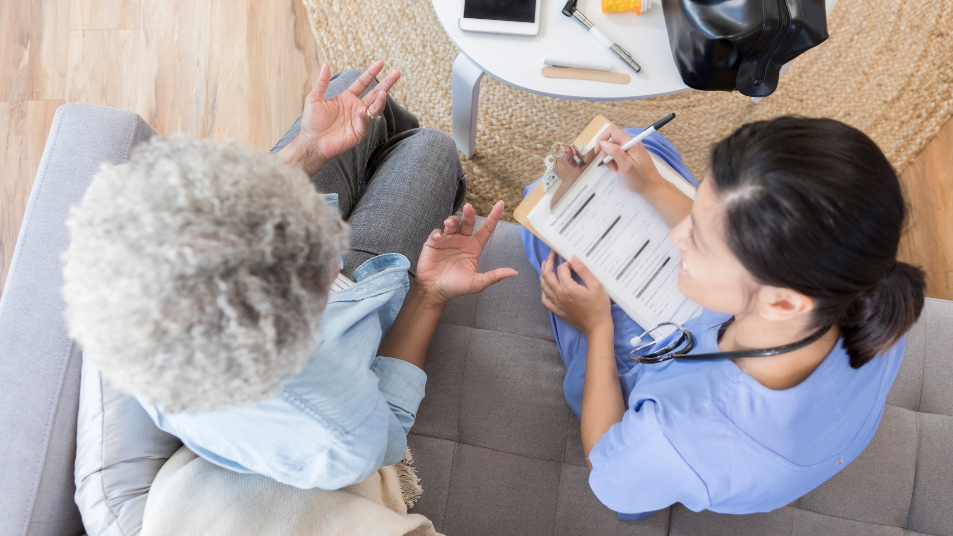 An older woman explains something to a nurse with a clipboard on a couch.
