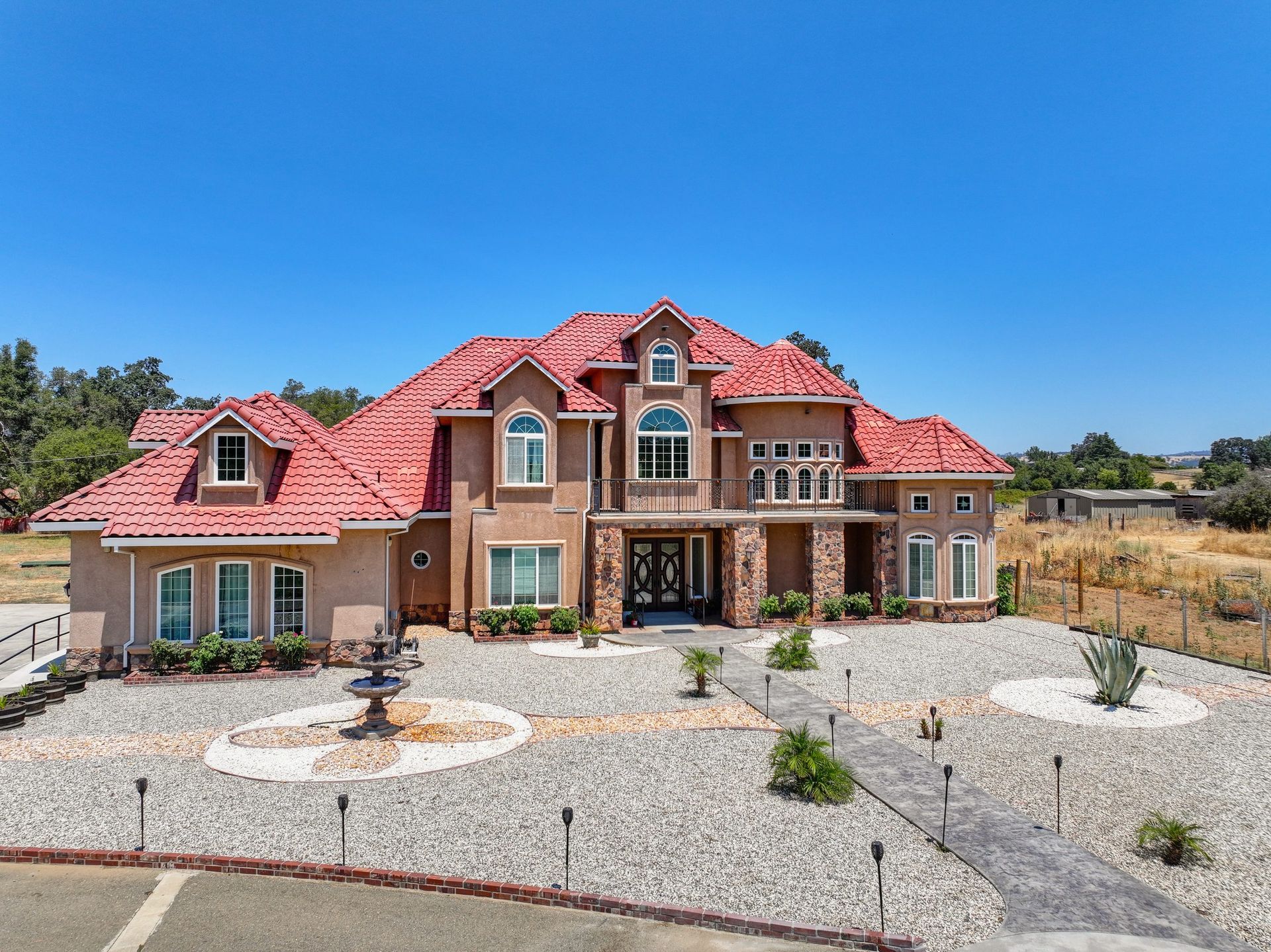 Two-story beige house with red tile roof, circular gravel driveway, fountain, and blue sky.
