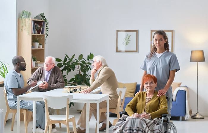 Care home: Elderly people at a table with a nurse, one woman in a wheelchair, a doctor talking to others.