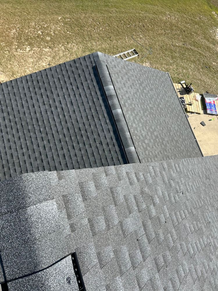 A gray shingled roof with a metal chimney pipe and cap, viewed from an elevated angle with a grassy lawn and trees behind.