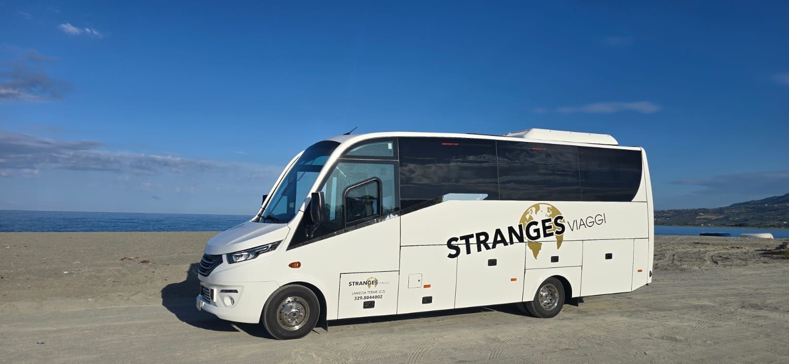 Autobus bianco con il logo "Stranges" parcheggiato su una spiaggia con il mare e il cielo sullo sfondo.