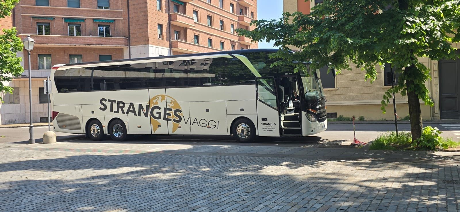 Un autobus turistico bianco con la scritta "Stranges Viaggi" è parcheggiato su una strada acciottolata accanto a un edificio.