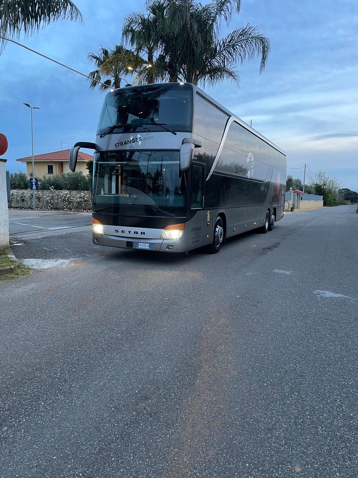 Un grande autobus a due piani color argento è parcheggiato su una strada asfaltata. È il crepuscolo e la scena è all'aperto.