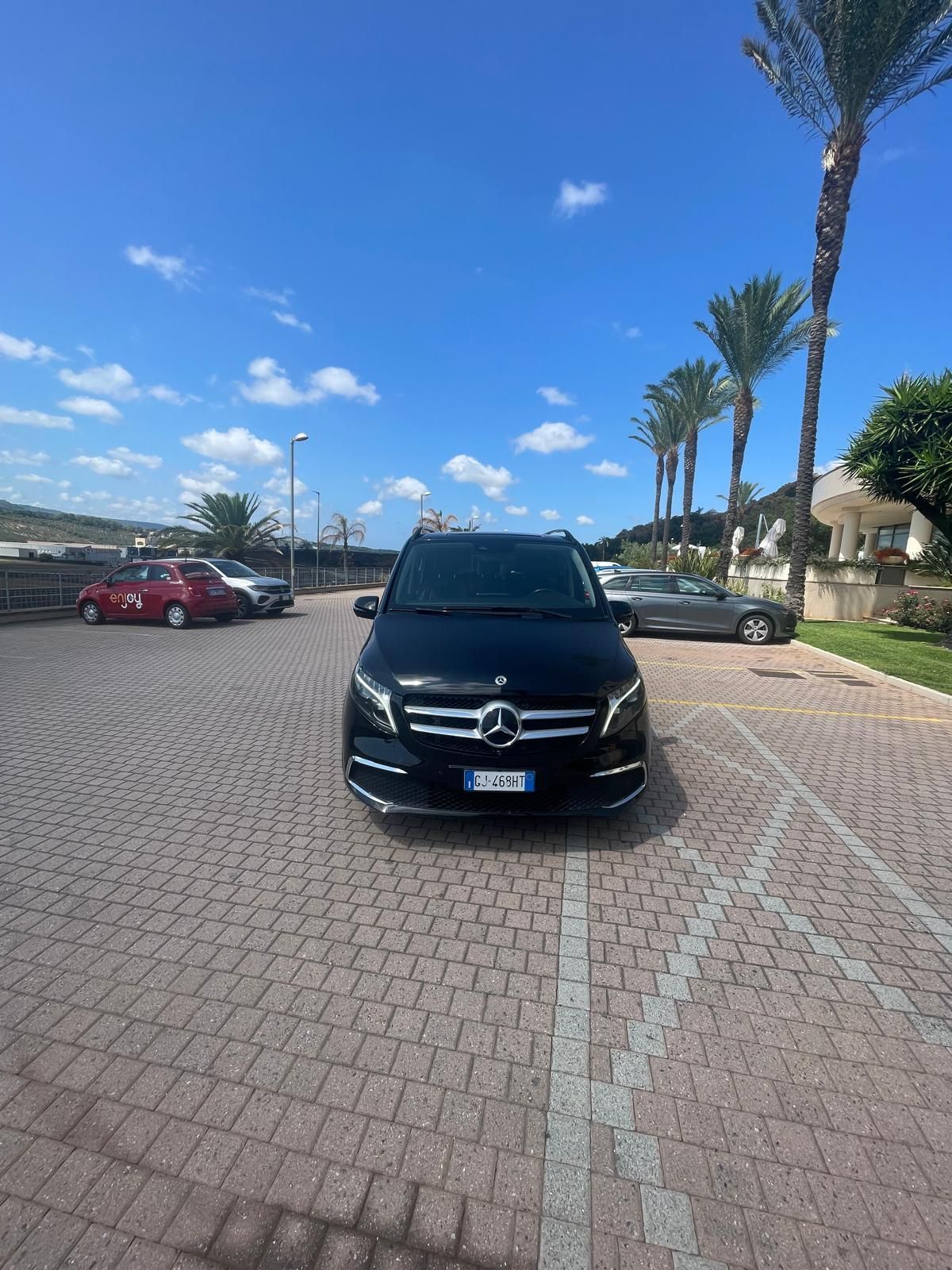 Un furgone Mercedes nero parcheggiato sul selciato sotto un cielo azzurro, vicino a palme e altre auto.