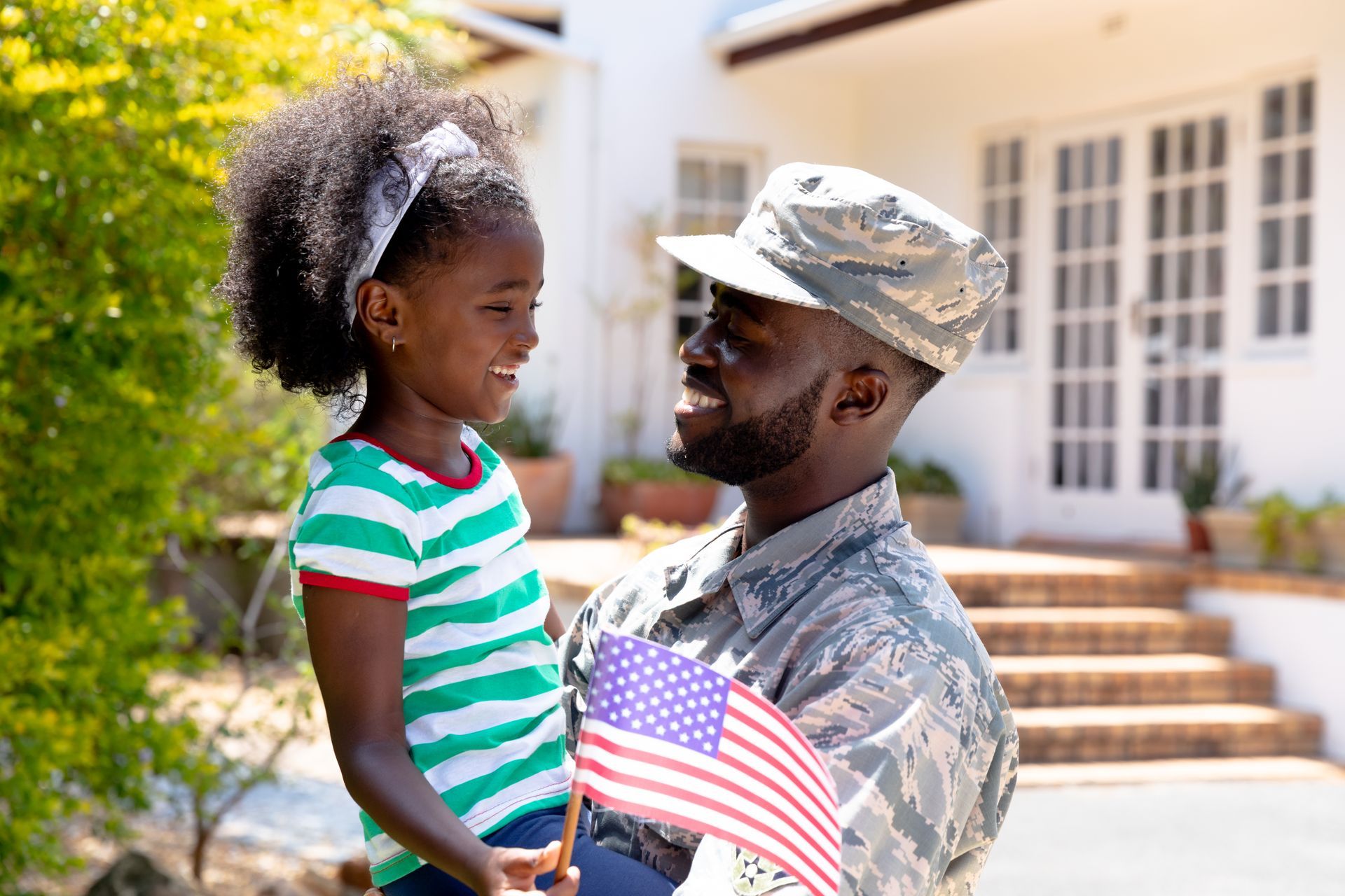 A man in a military uniform is holding an American flag and a little girl.