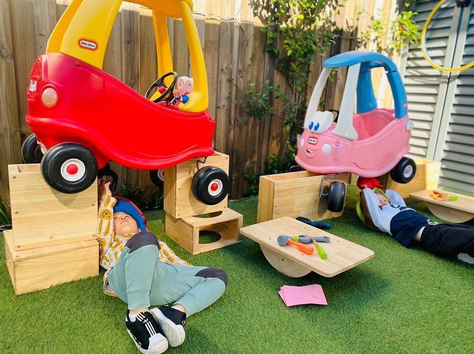 Two children lie on the grass under toy cars elevated on wooden crates in an outdoor play area.