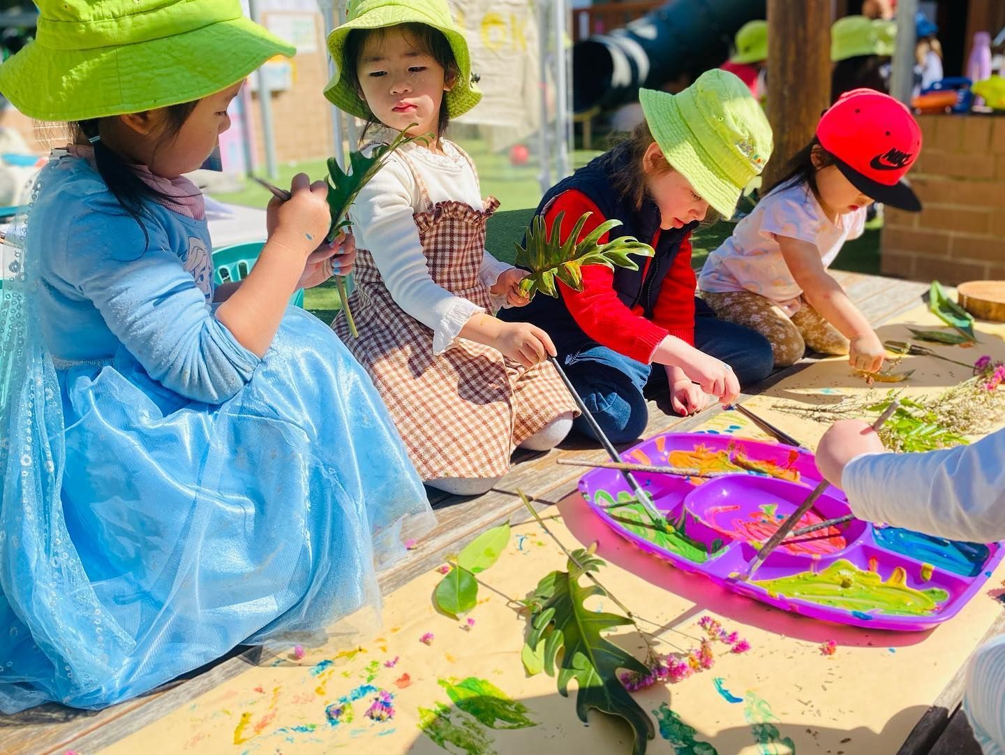 Four children wearing hats are outdoors, painting leaves with colorful paints on a paper-covered table.