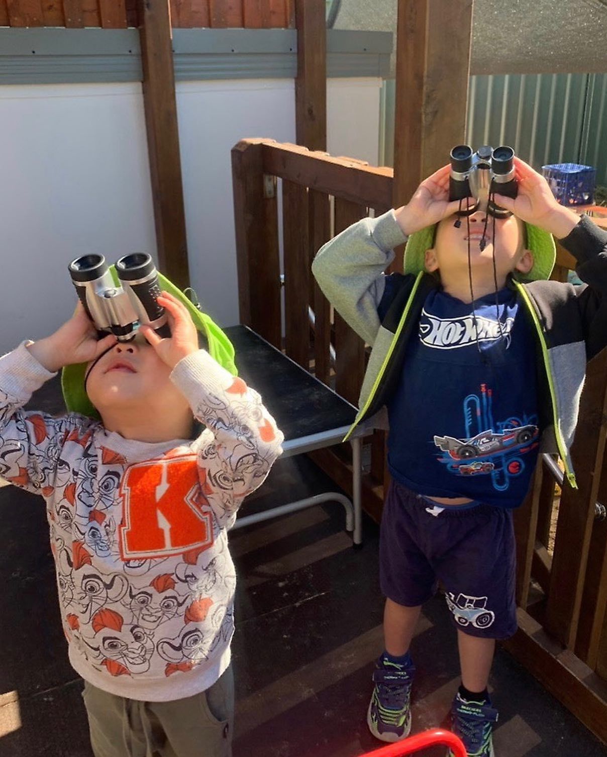 Two children wearing green hats stand outside looking up at the sky through toy binoculars.