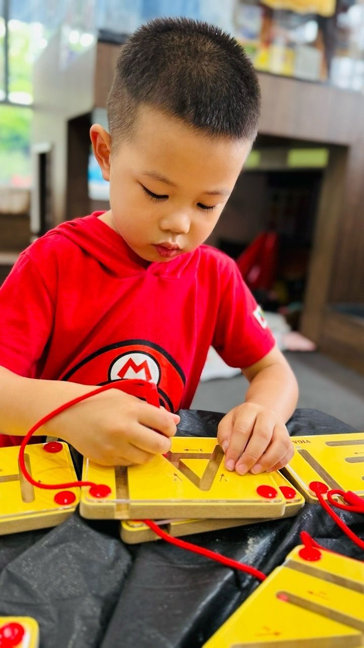 A child in a red shirt traces the letter 'A' on a yellow wooden board with a red string toy.