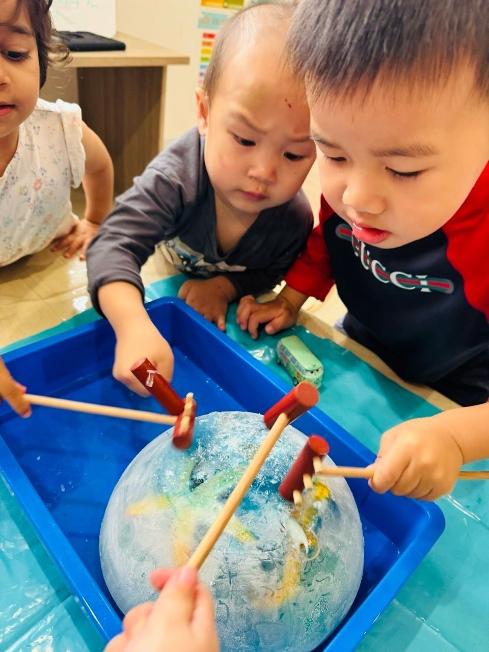 Three children use small mallets to chip at a large, round block of ice inside a blue plastic tray.