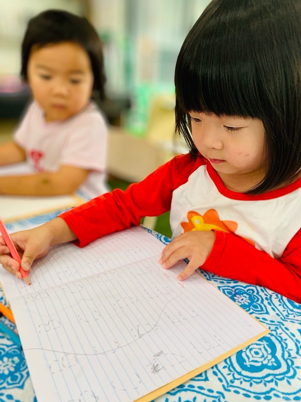 Two children sit at a table with a patterned cloth, focusing on drawing in lined notebooks.