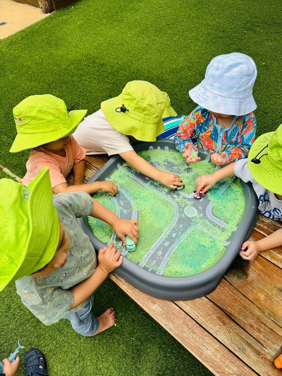 Four children wearing hats play together around a plastic table featuring a toy road map filled with green filler.
