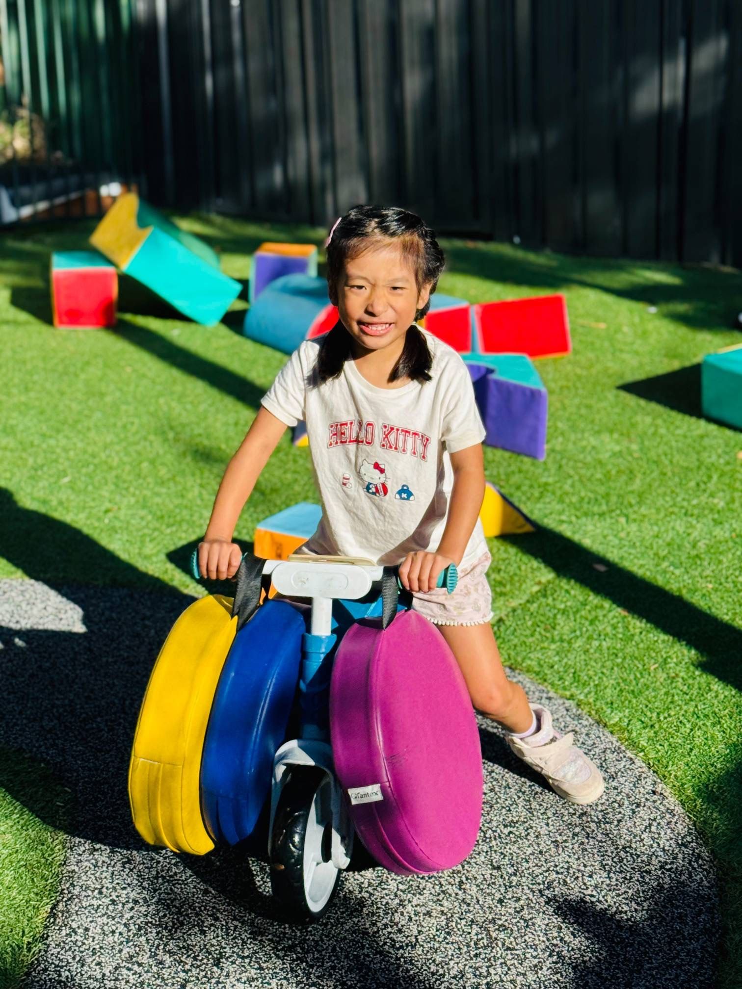 A smiling child sits on a three-wheeled toy bike, featuring large yellow, blue, and purple circular pads, in a park.