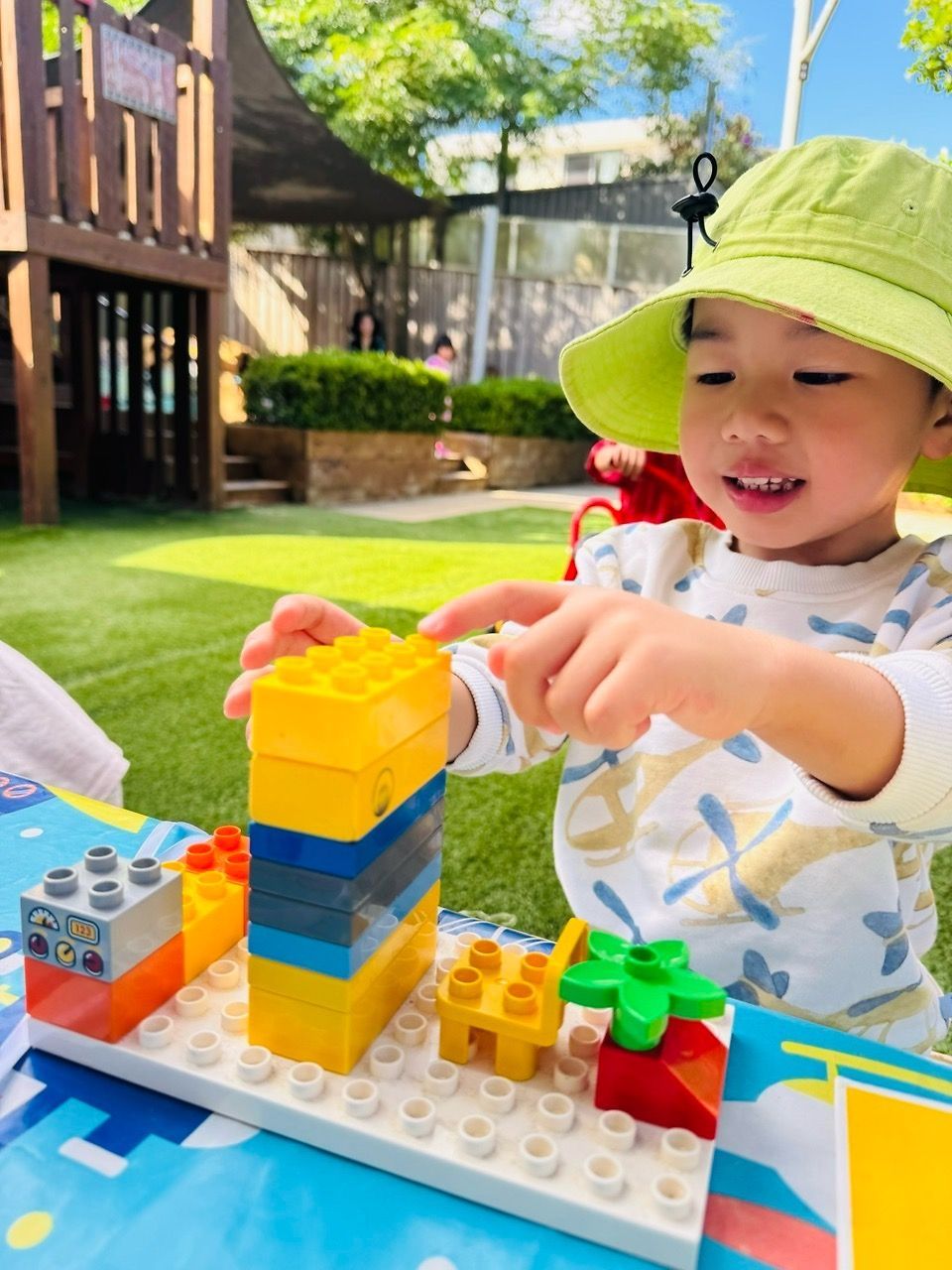 A smiling child in a lime green bucket hat plays with colorful plastic building blocks outdoors on a sunny day.