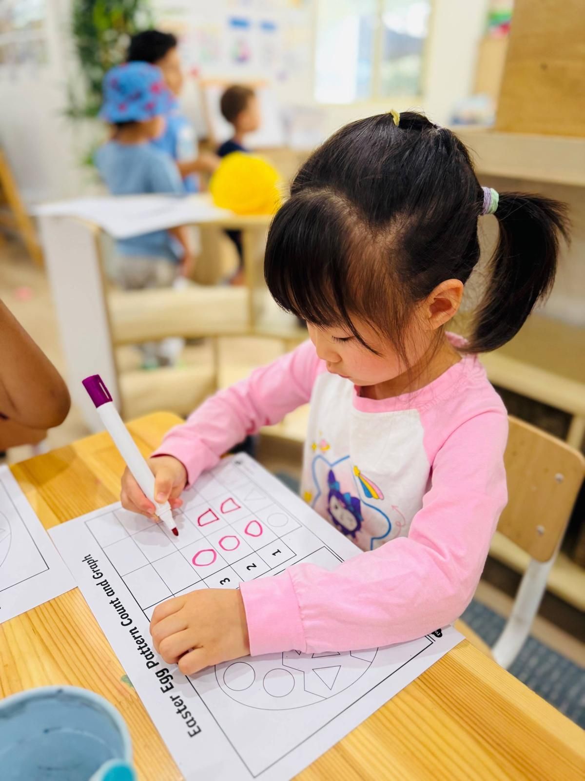 A child in a pink shirt sits at a wooden table, focused on filling out an