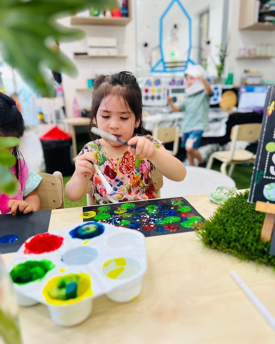 A child painting with vibrant colors on a black surface, using a palette in a classroom with a rocket mural background.