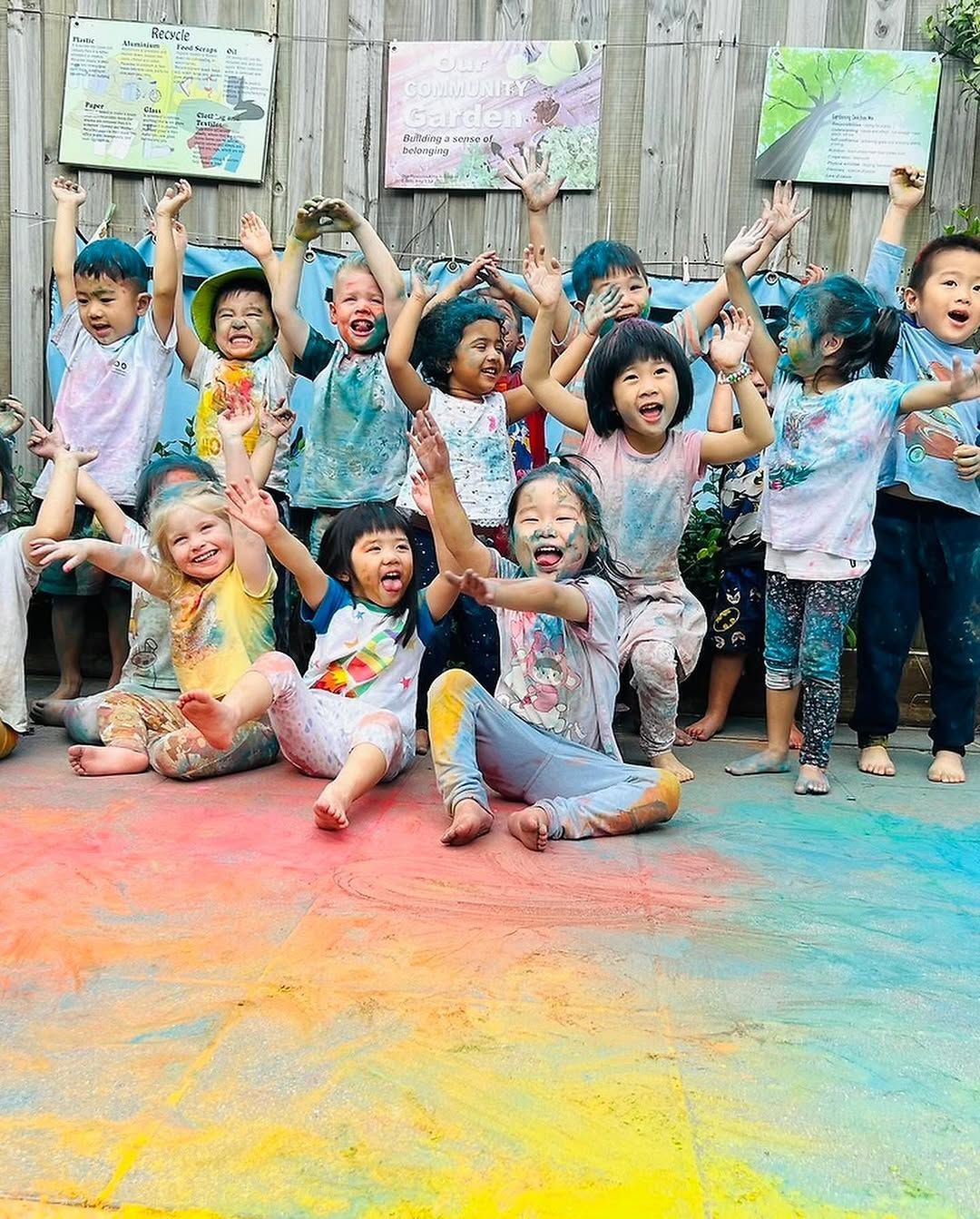 A large group of children covered in colorful powder celebrate outdoors, smiling and throwing colors in the air.