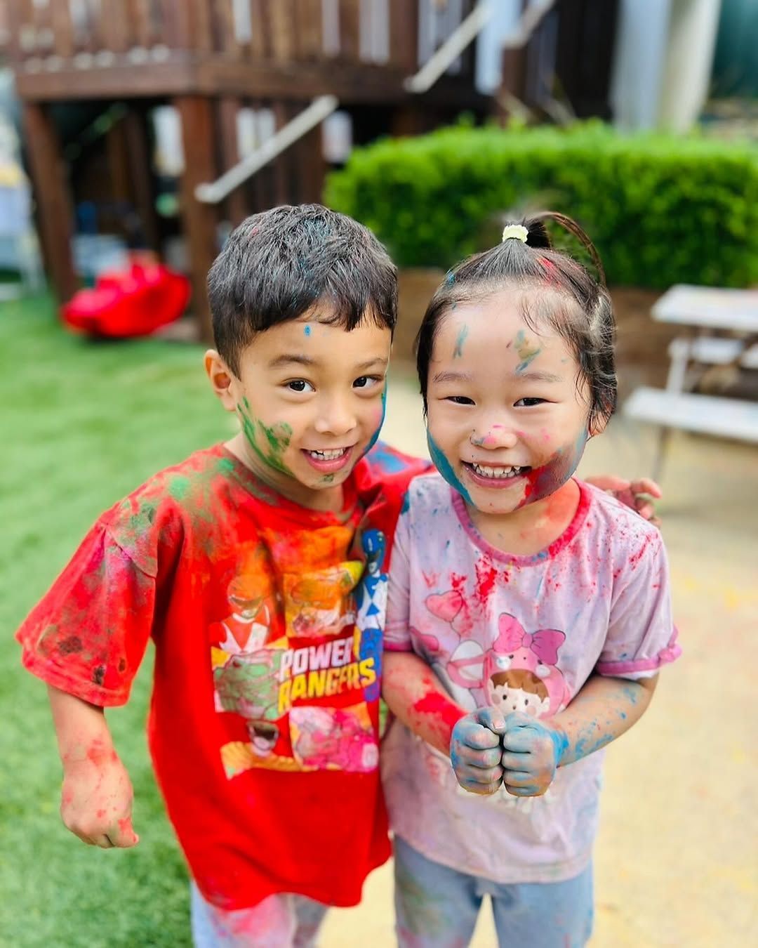 Two smiling children covered in colorful paint standing side-by-side outdoors on a sunny day.