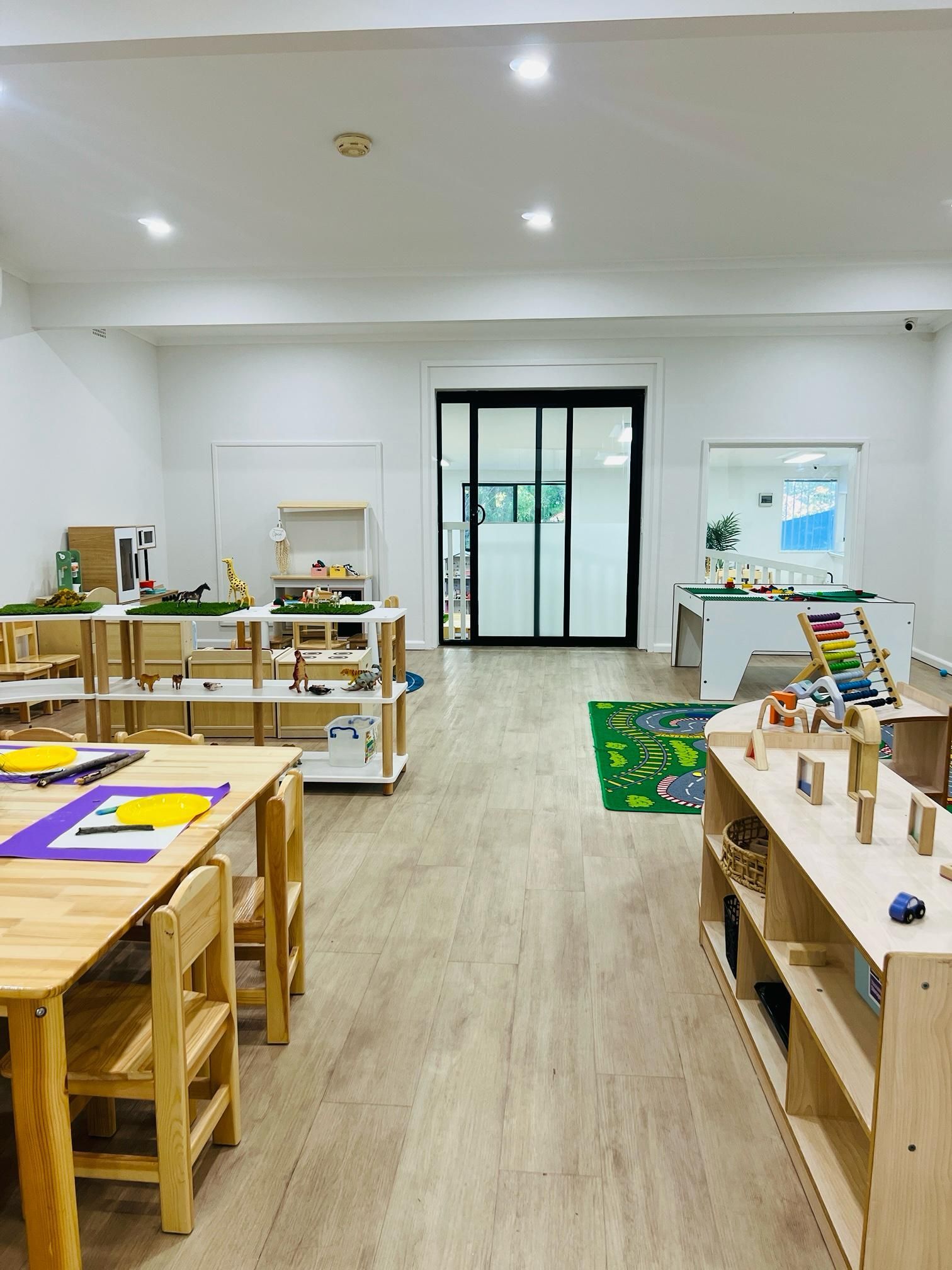 A brightly lit, modern preschool classroom with wooden tables, child-sized chairs, and open shelving filled with toys.