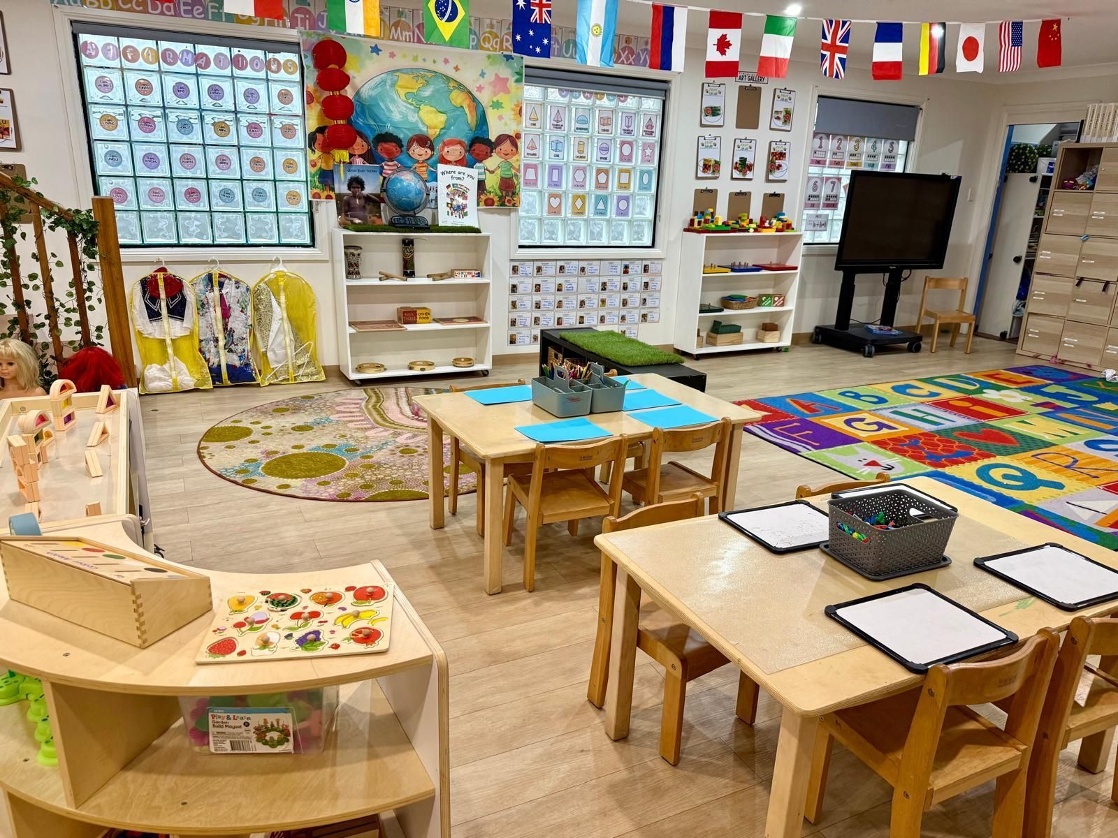 A bright preschool classroom with tables, chairs, a rug, educational posters, and international flags hanging overhead.
