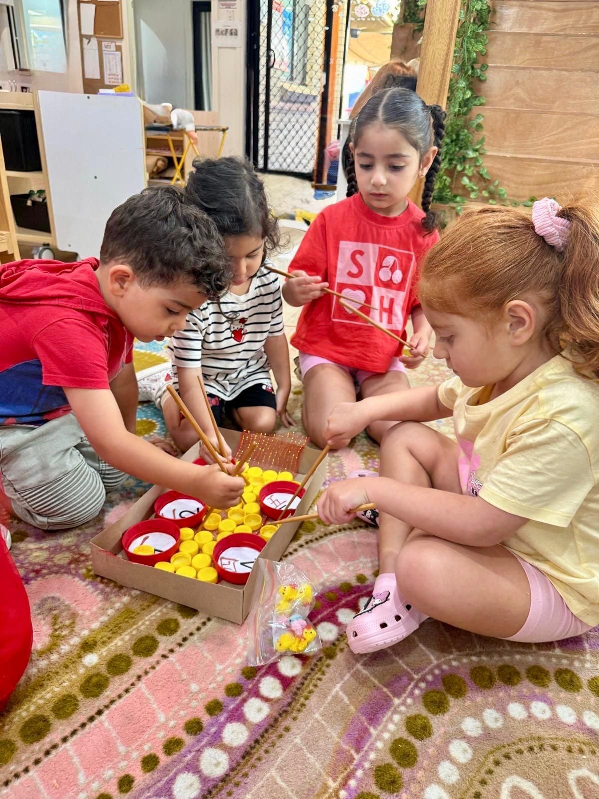Four children sit on a patterned rug, using small tools to transfer yellow pom-poms into colorful bowls in a cardboard tray.