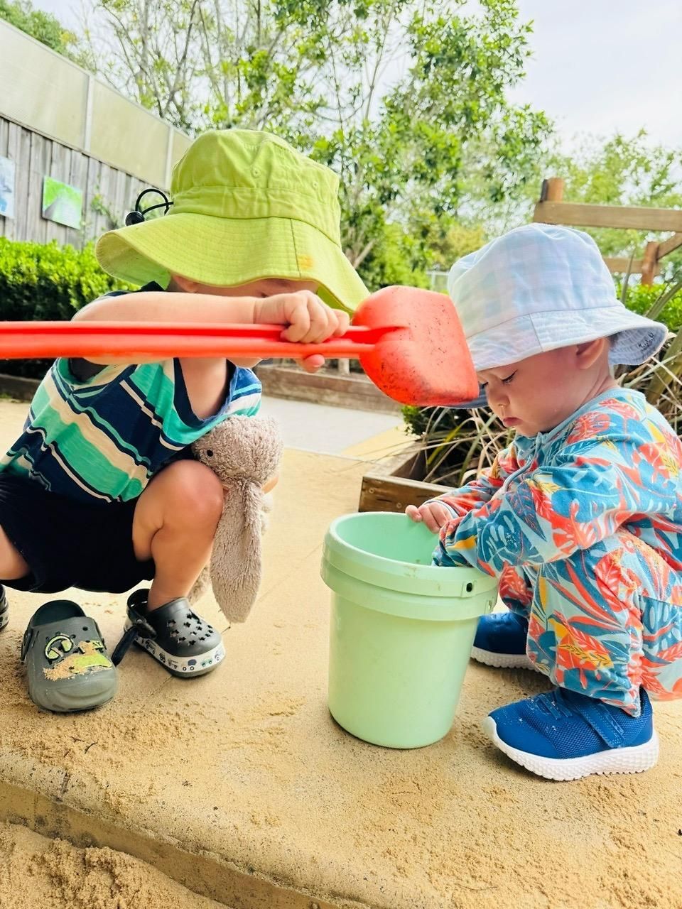 Two toddlers in sun hats play with a plastic shovel and pale green bucket in a sandy outdoor area.