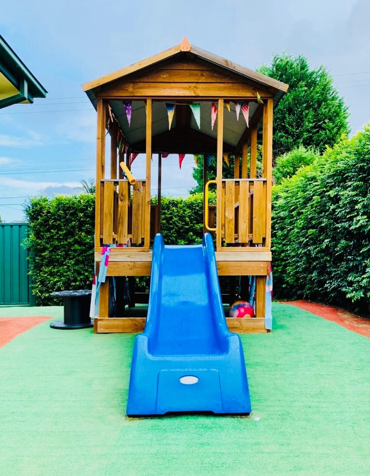 A wooden playhouse with a blue plastic slide in a backyard with green hedges and synthetic grass.