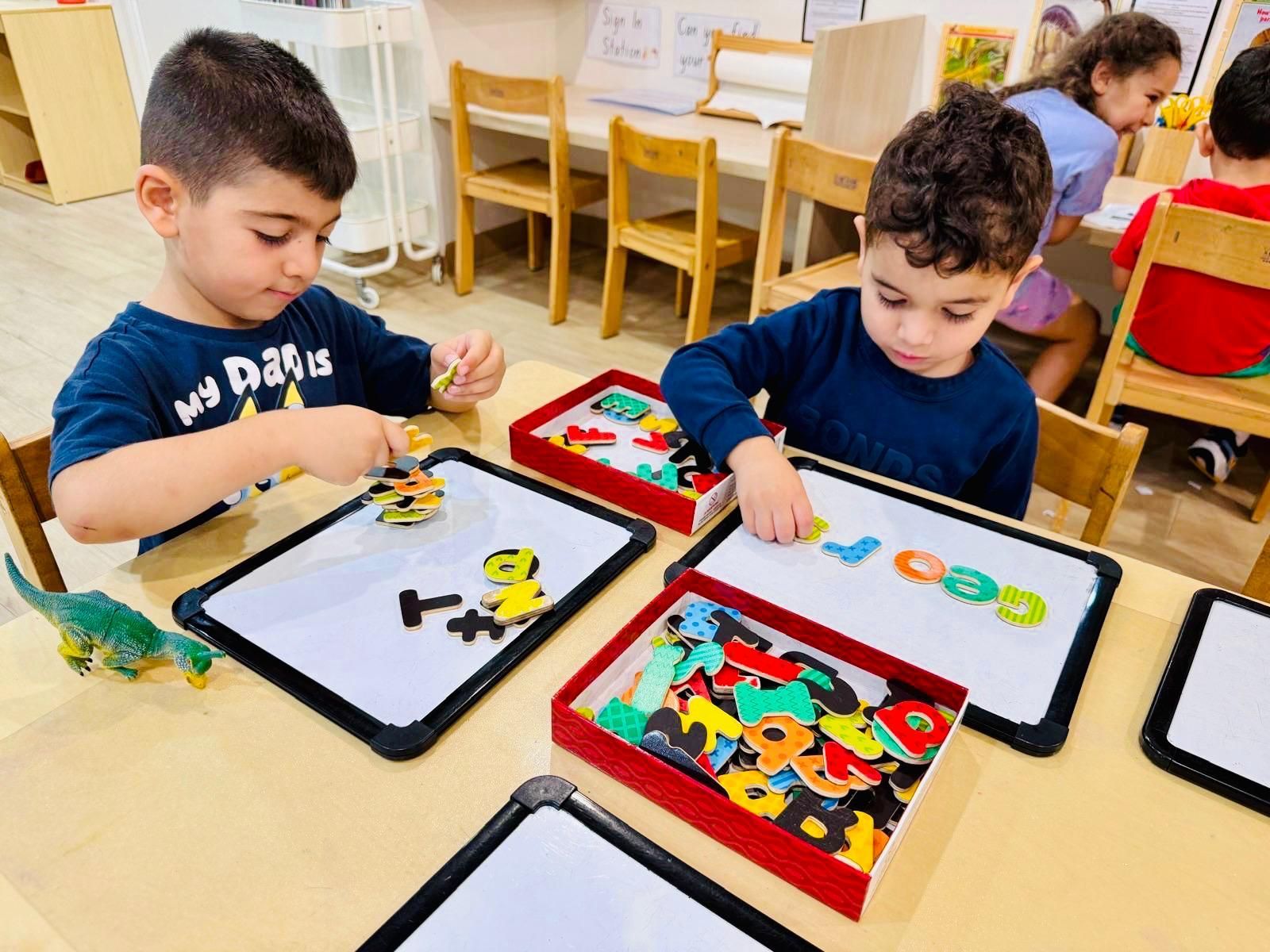 Two children in a classroom work with colorful magnetic letters on small whiteboards, with a box of letters nearby.