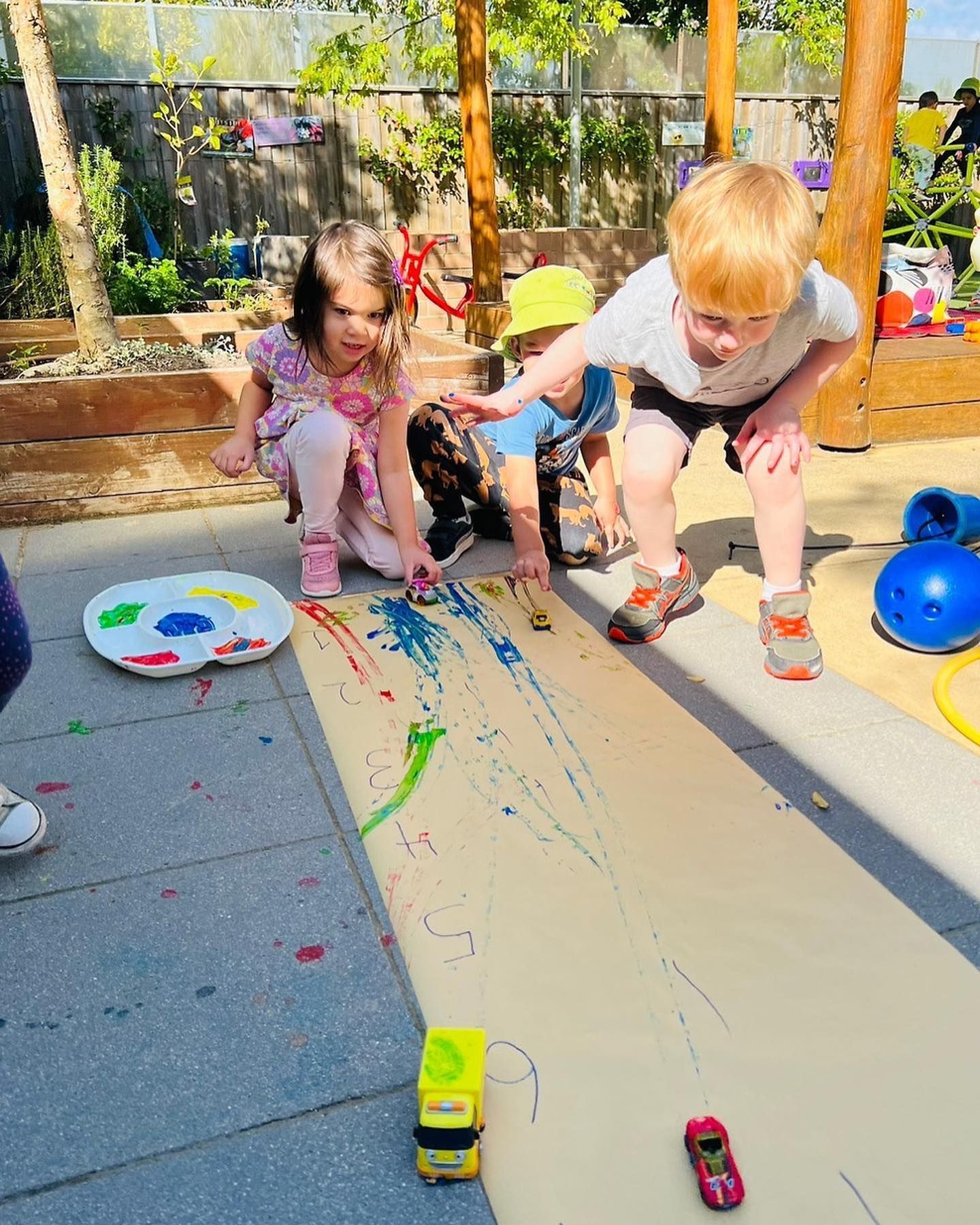Children paint on a long paper strip using toy cars as brushes in an outdoor play area.