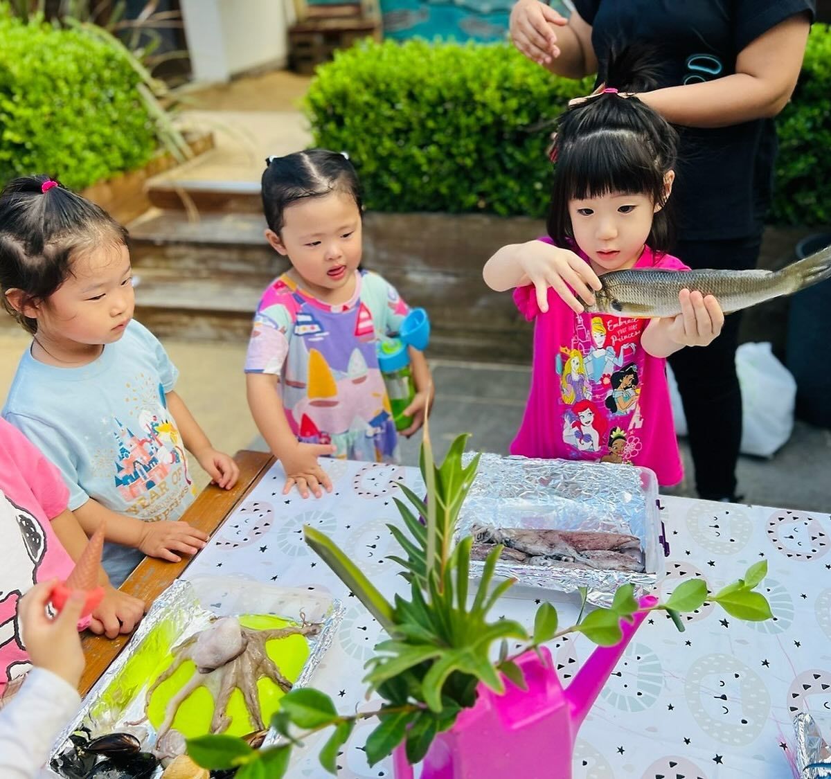 Children around a table inspect a raw fish and an octopus laid out on foil, in an outdoor setting.