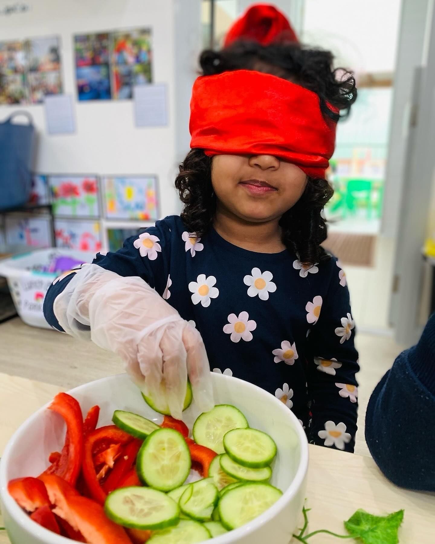 A person wearing a blindfold and a glove selects a cucumber slice from a bowl filled with sliced peppers and cucumbers.