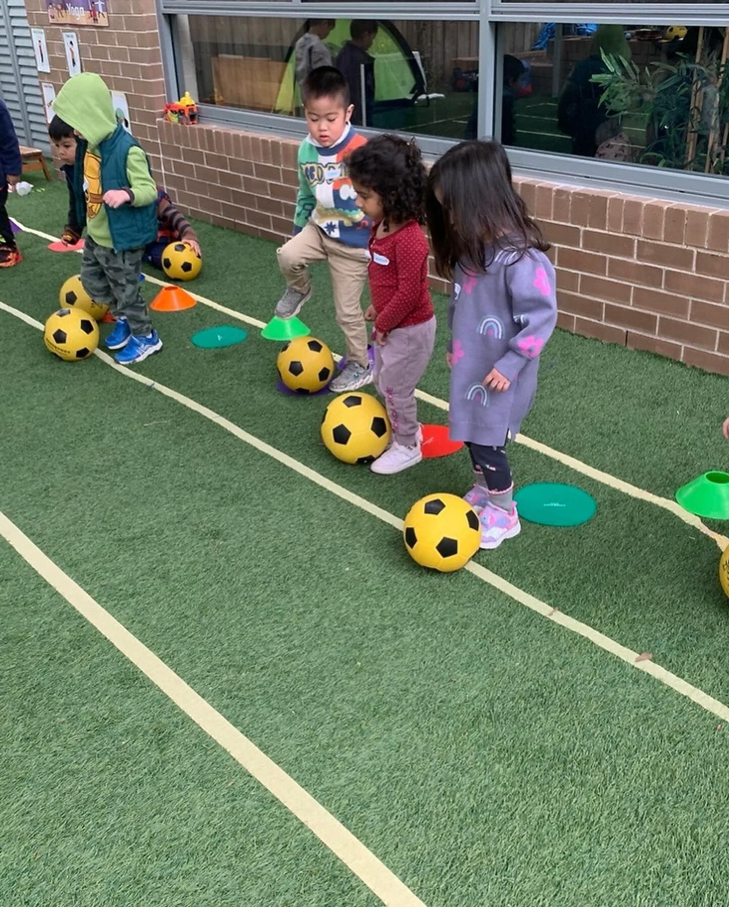 Children in active wear participate in a soccer drill, practicing ball control on an outdoor synthetic grass court.