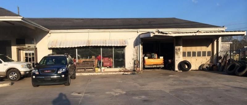 A car repair shop with two cars parked in front. Shop has open garage door and tires stacked outside.
