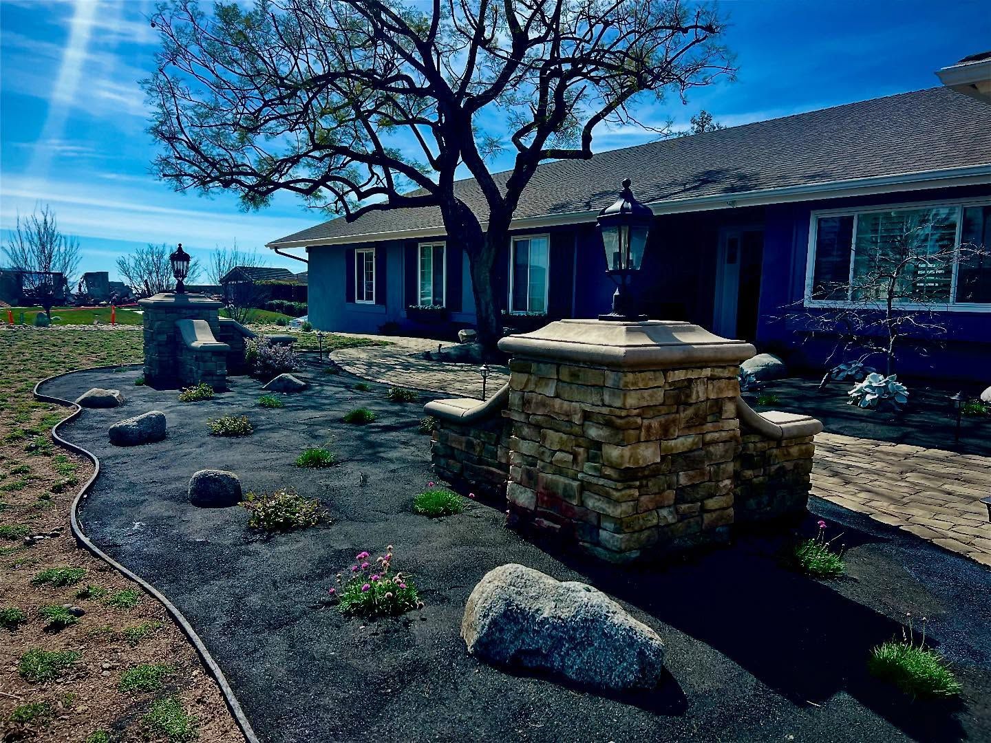 A blue house with stone pillars, landscaping, and a large tree under a bright blue sky.