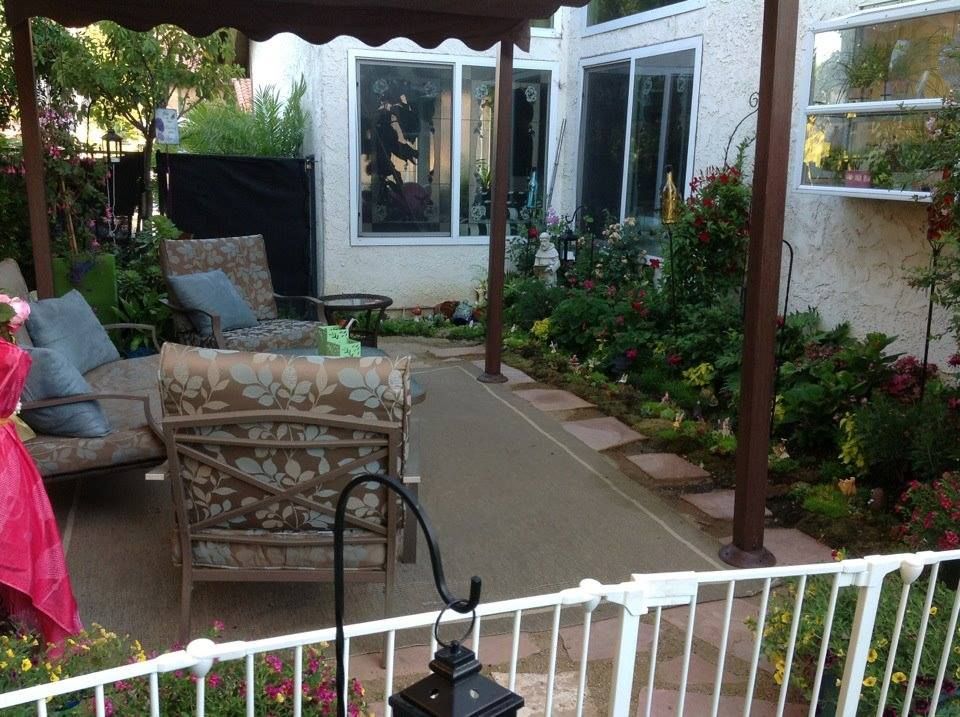 Patio with seating under a canopy, a concrete path, and a garden with flowers along a white-walled building.
