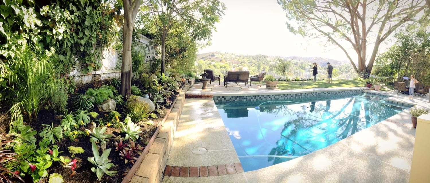 A lush garden surrounds a sparkling pool. Two people stand in the distance near patio furniture and a scenic view.
