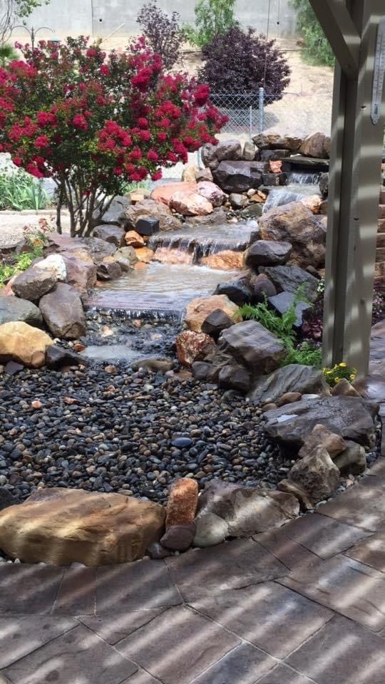A backyard water feature with a cascading stream surrounded by rocks and plants. A pergola is in the foreground.