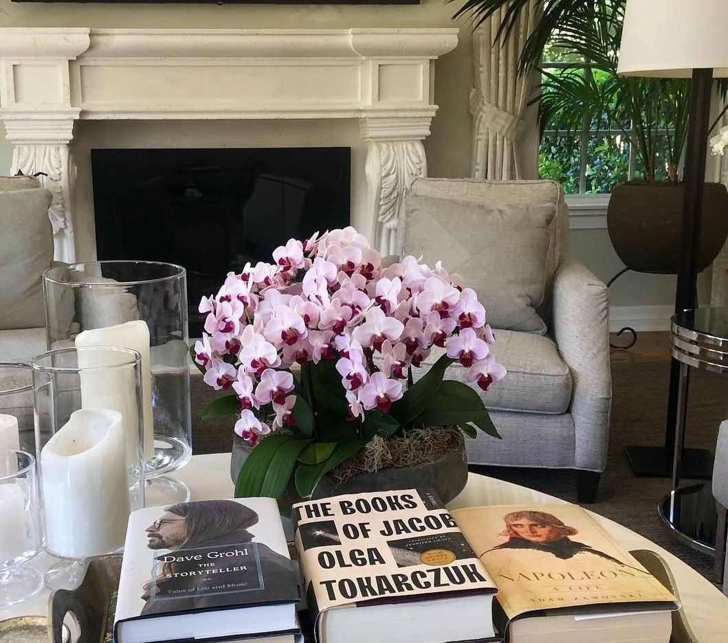 Pink orchids on a coffee table with books and candles. Behind are a fireplace, chairs, and a window.