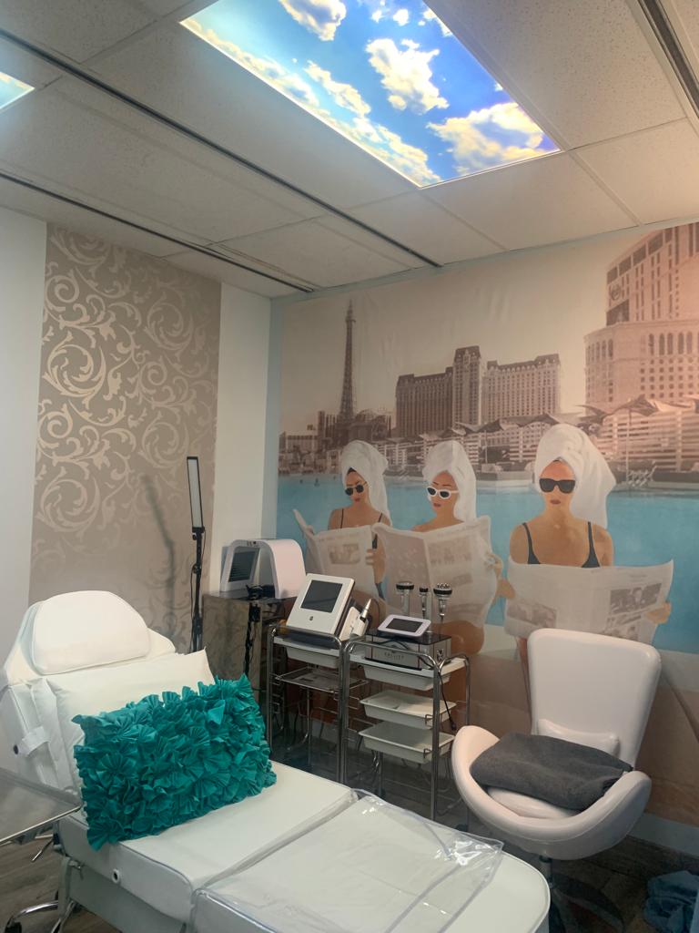 A room with a bed , chair , and a picture of two women in towels on the wall.