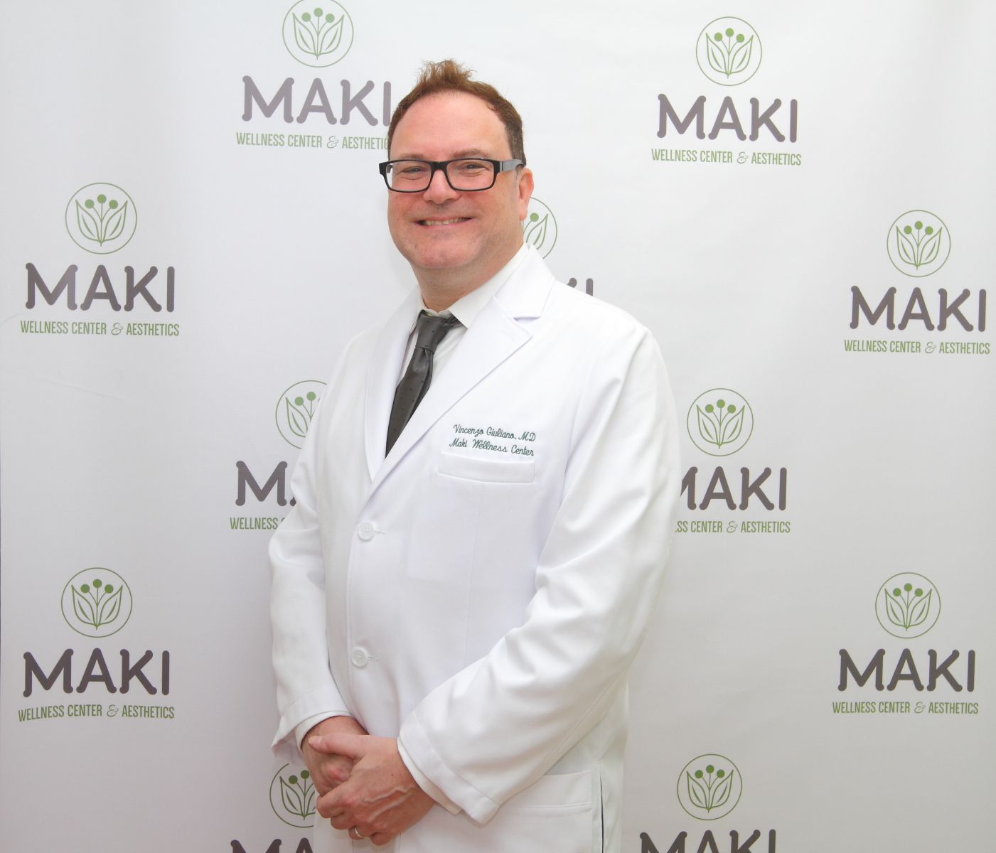 A man in a white coat and tie is standing in front of a wall that says maki