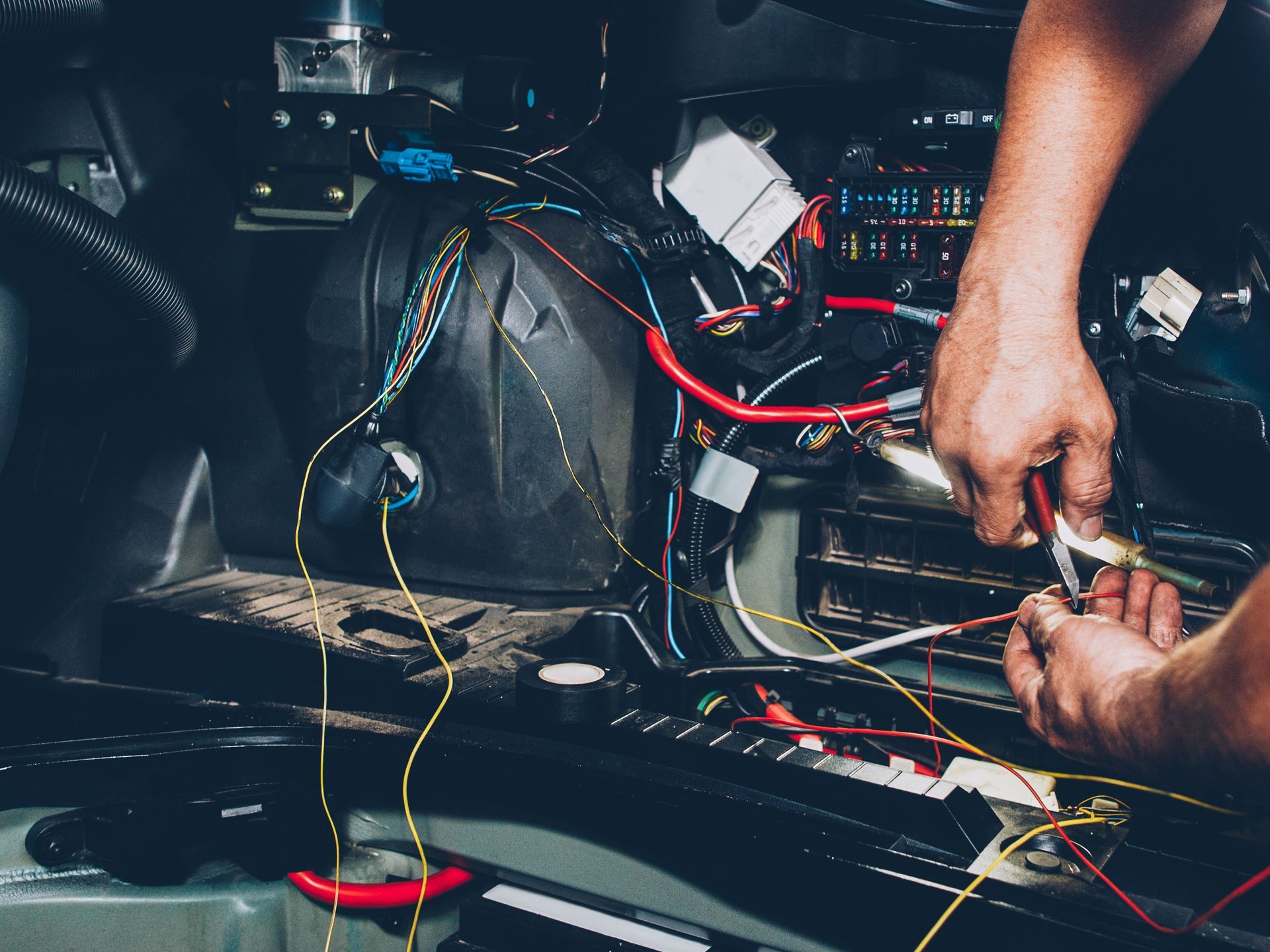 A person’s hands using pliers to wire electrical components inside the dark engine compartment of a vehicle.