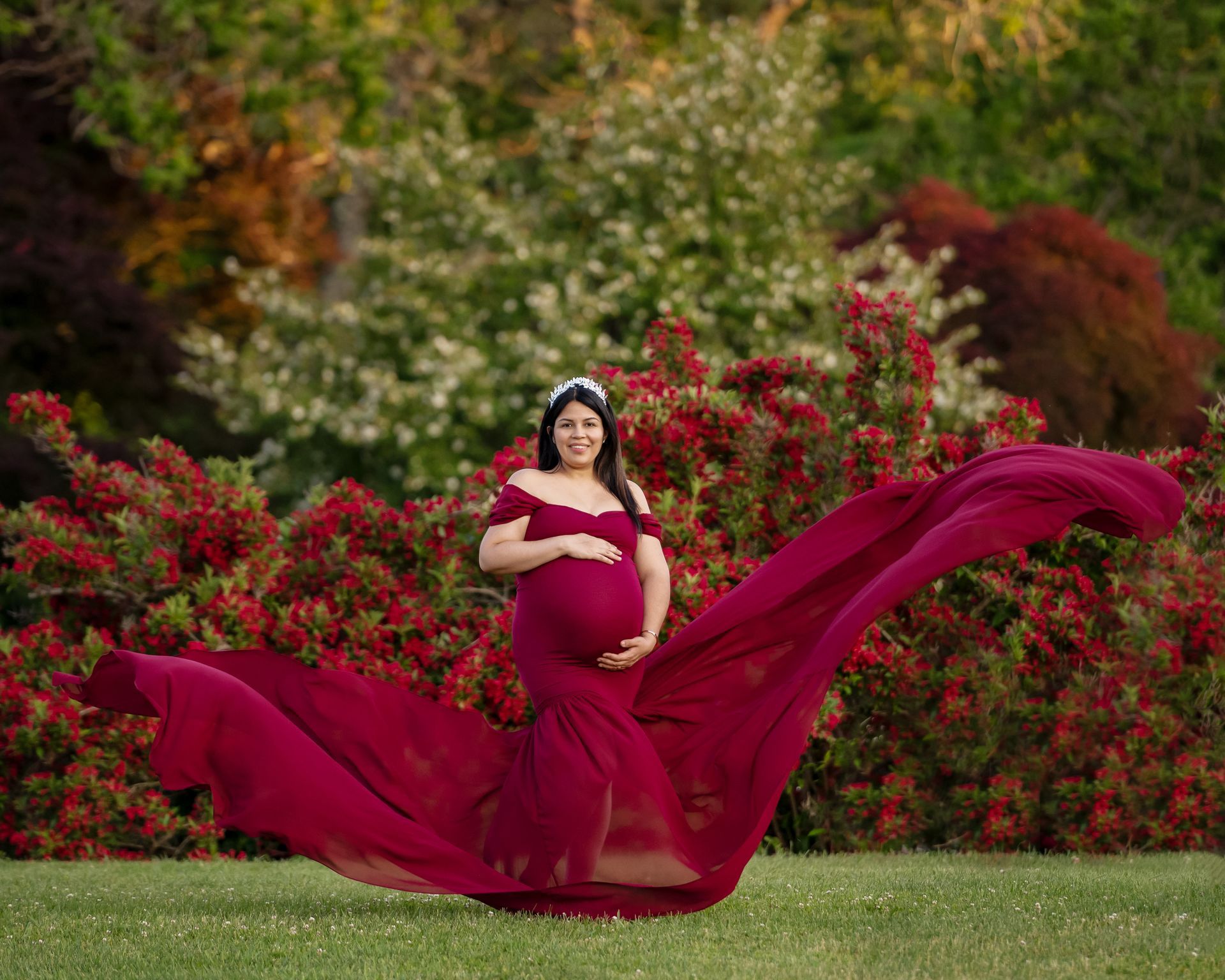 a pregnant woman in a long red dress is standing in front of a bush of red flowers .