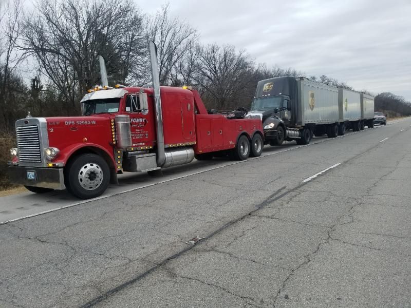 Red tow truck towing a UPS semi-truck with trailers on a highway. Cloudy day.